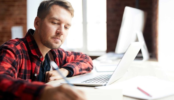 Junger Mann sitzt vor Laptop und macht sich Notizen auf seinem Block